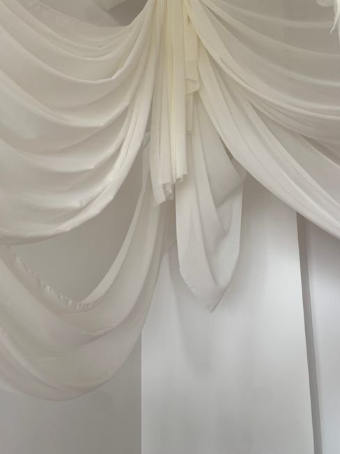 Close-up of elegant, white, fabric curtains with multiple pleats and drapes, arranged in a layered style, hanging from a curtain rod in a well-lit interior space. The fabric appears clean and well-mai