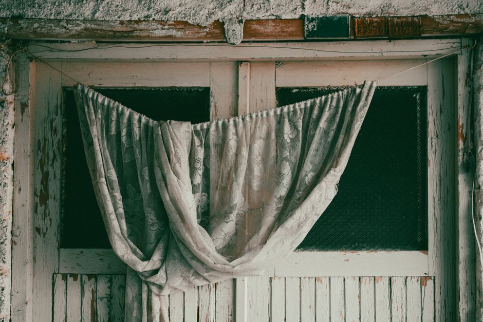 Close-up of elegant, white, fabric curtains with multiple pleats and drapes, arranged in a layered style, hanging from a curtain rod in a well-lit interior space. The fabric appears clean and well-mai