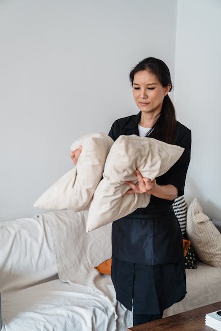 A close-up view of a person using a handheld electric upholstery cleaner with a round brush attachment on a dark gray fabric sofa in a living room. The individual, wearing gloves and dark clothing, pr